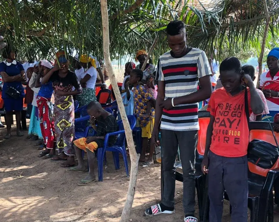 Believers standing in prayer at an outdoor gathering under palm leaves