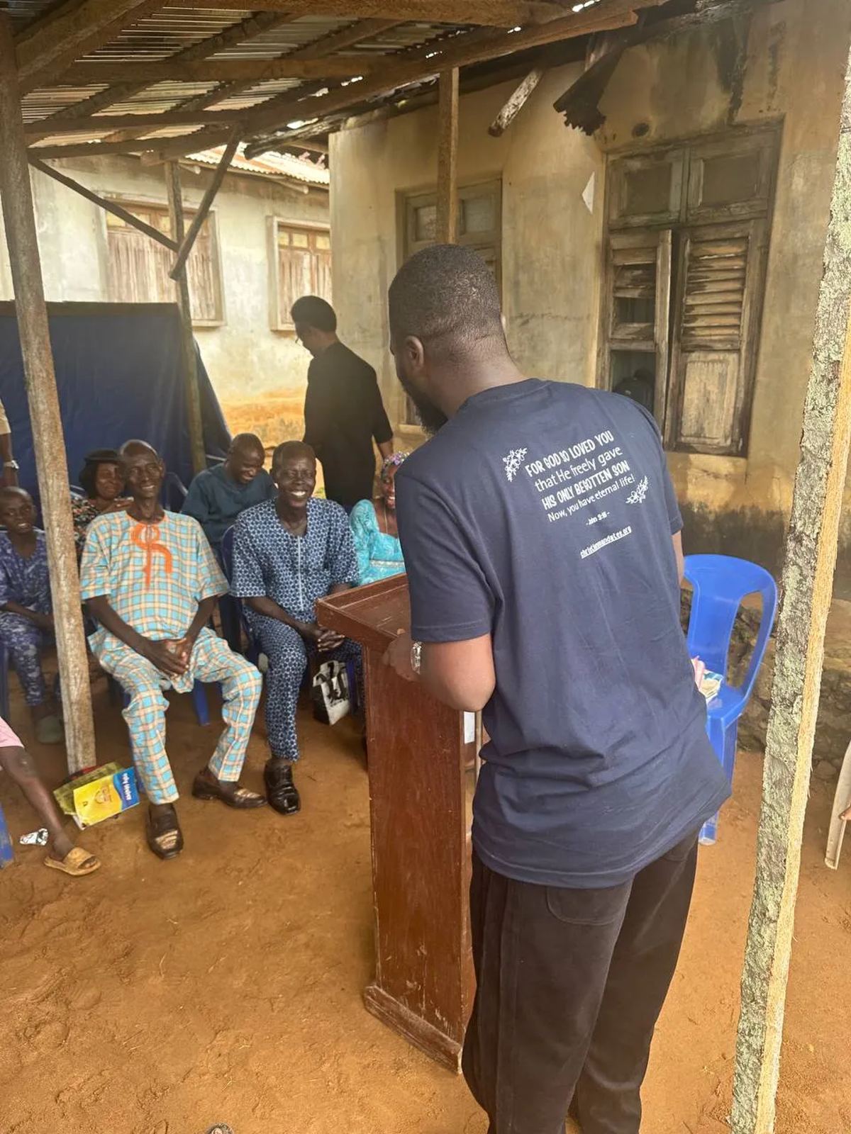 A team member at a lectern addressing people seated under a canopy