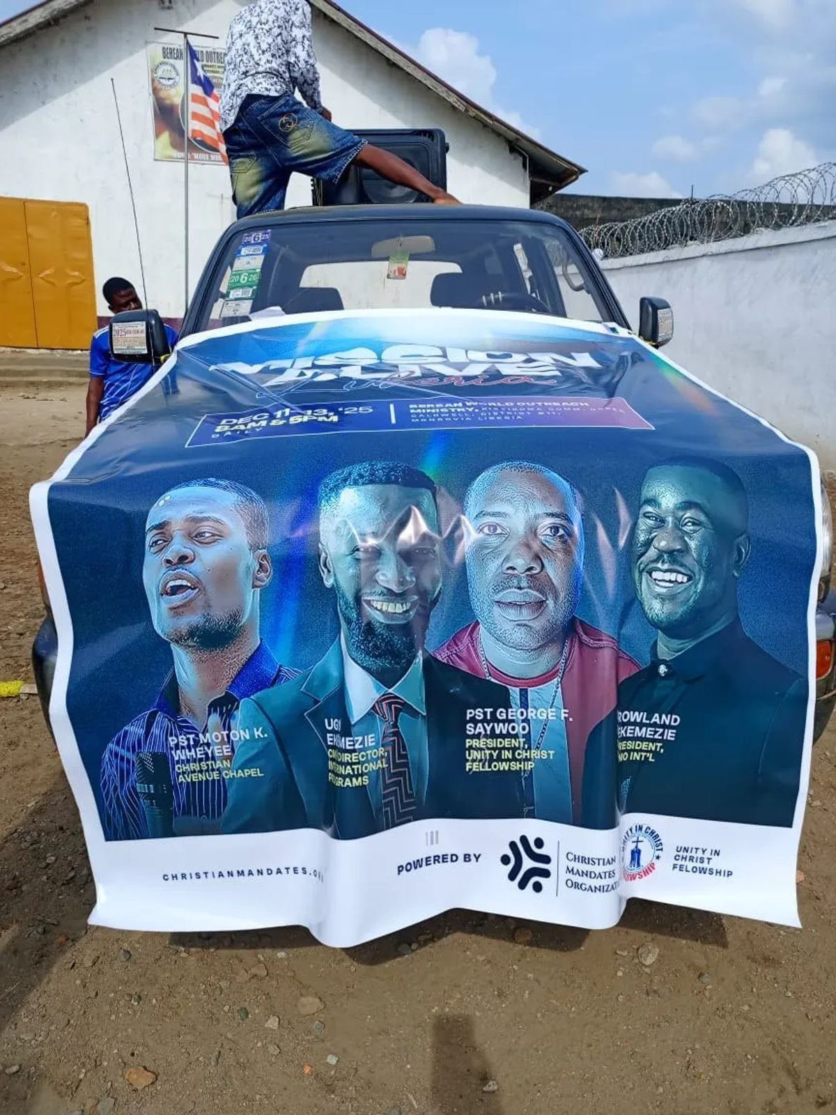 Mission Alive Liberia 2.0 banner with the four speakers' portraits, draped over the front of a car