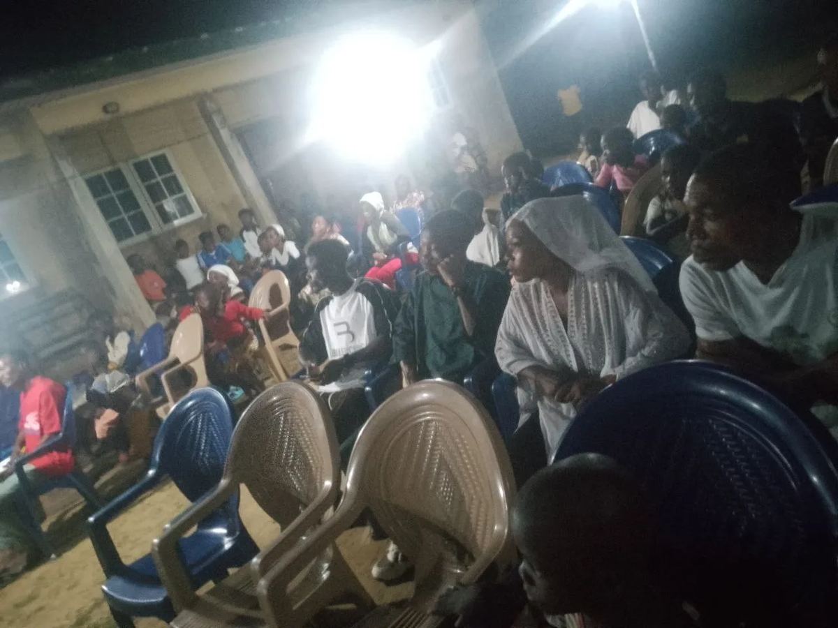 People seated on plastic chairs under floodlights at the night meeting