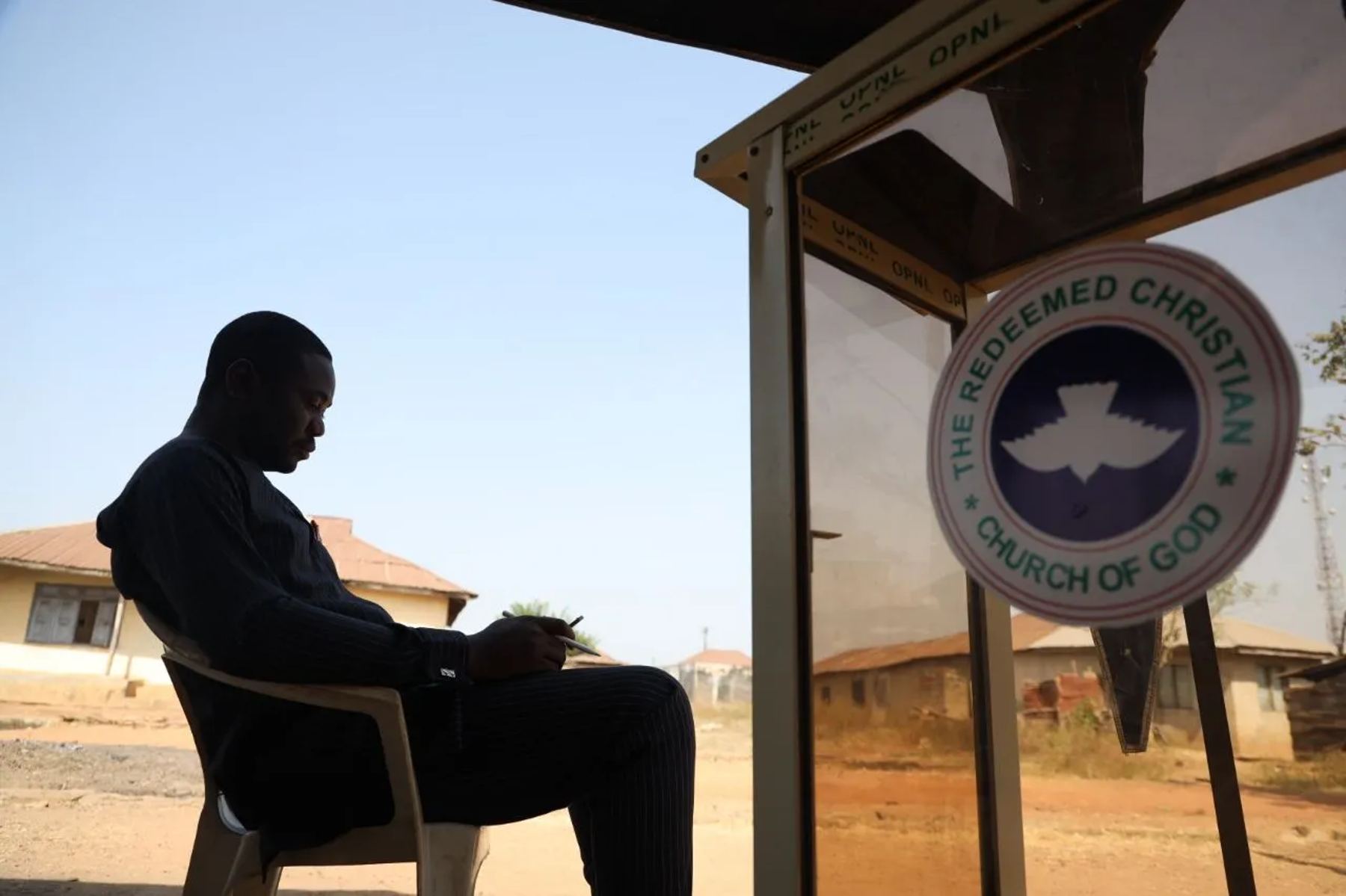 A man reading in the shade beside a Redeemed Christian Church of God sign