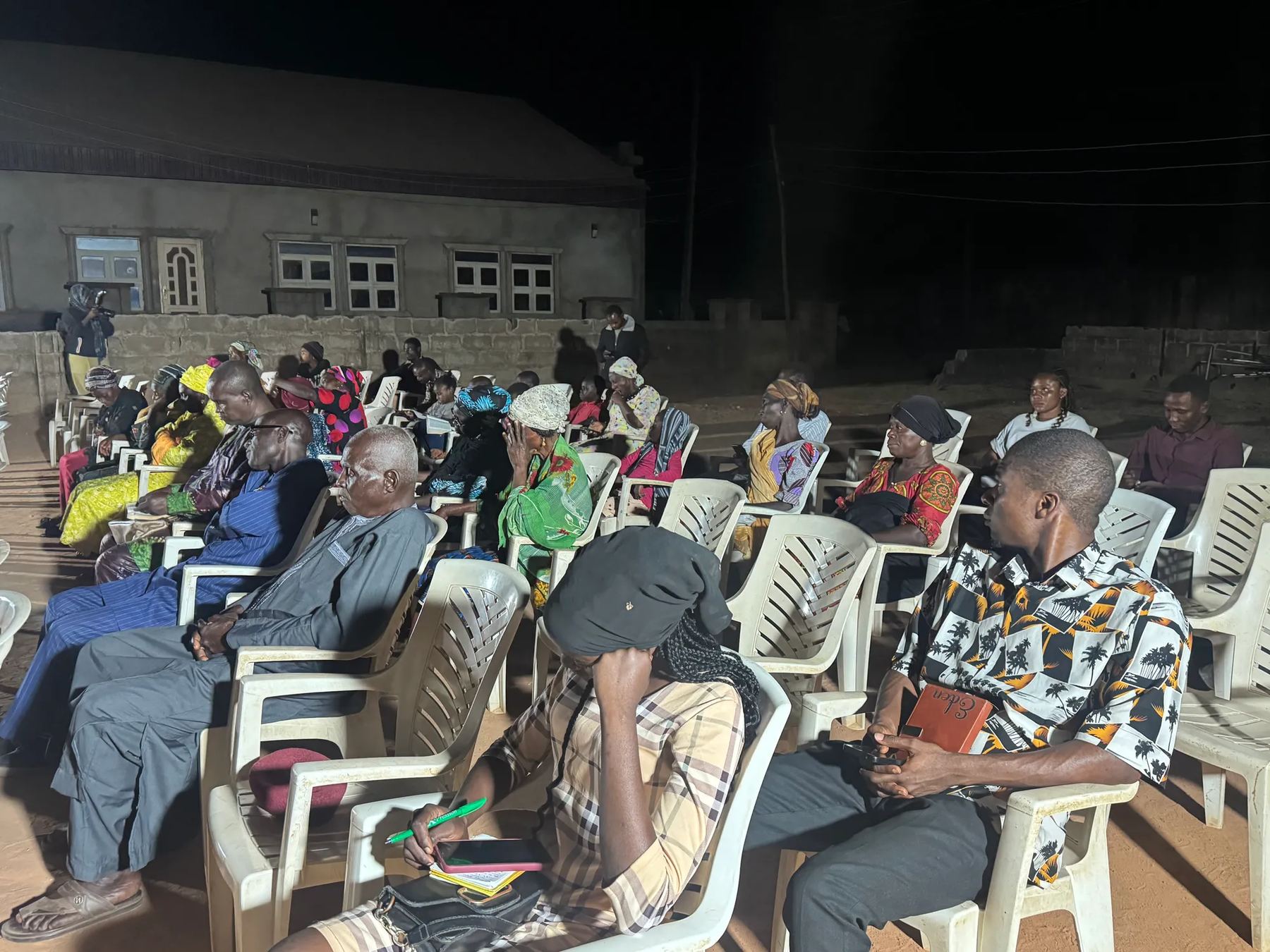 People seated on plastic chairs at an evening open-air meeting