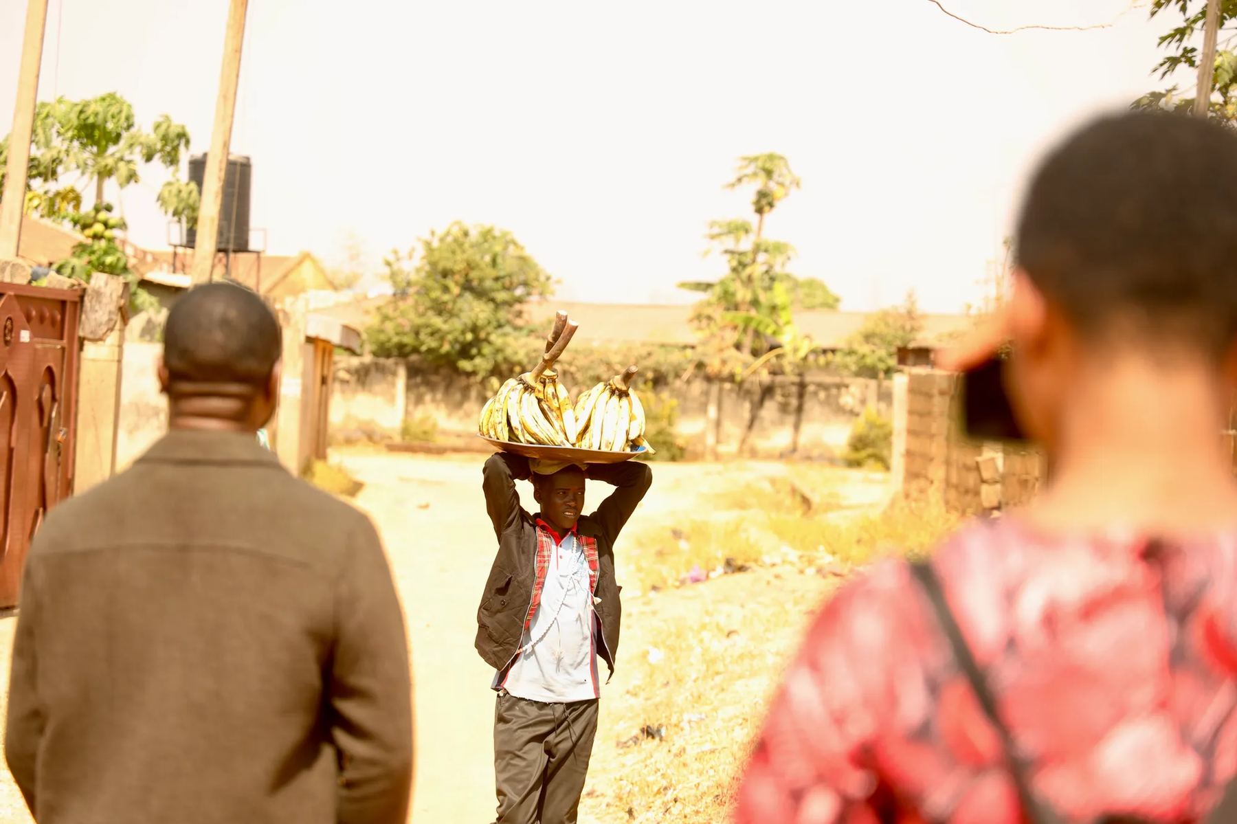 A woman carrying a tray on her head along a dirt road, seen past two team members