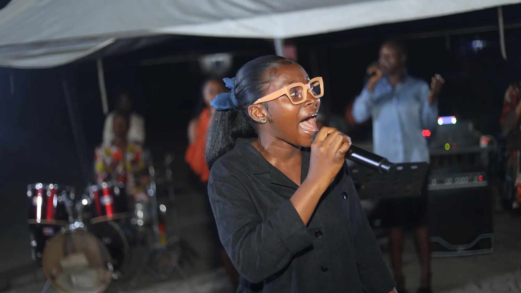A woman in glasses singing with a microphone at the evening crusade, with the band behind