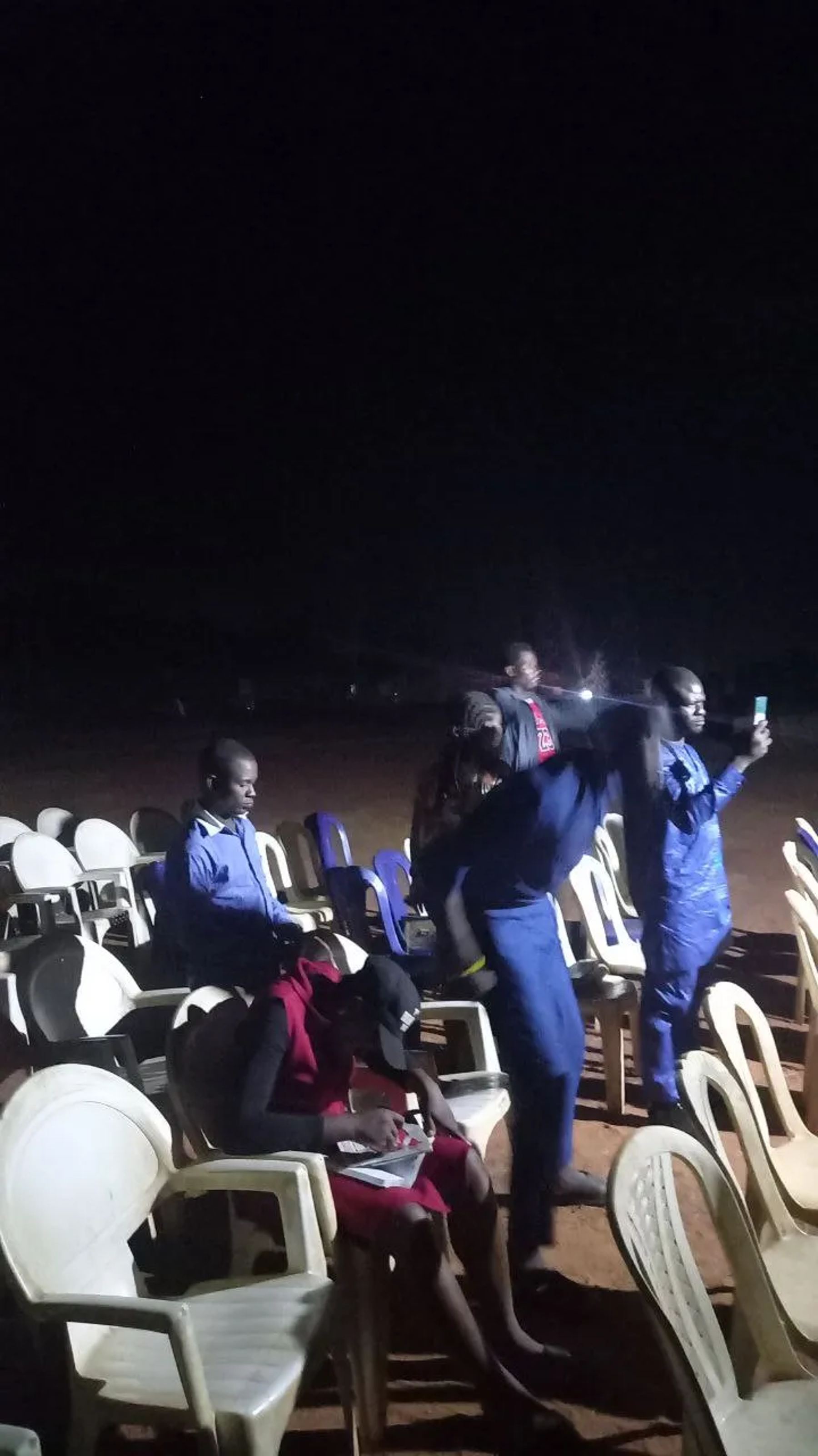 People seated on plastic chairs in an open field at night