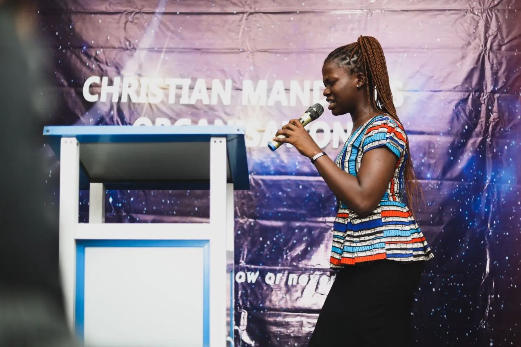 A woman speaking with a microphone beside a lectern, in front of a Christian Mandates Organisation banner