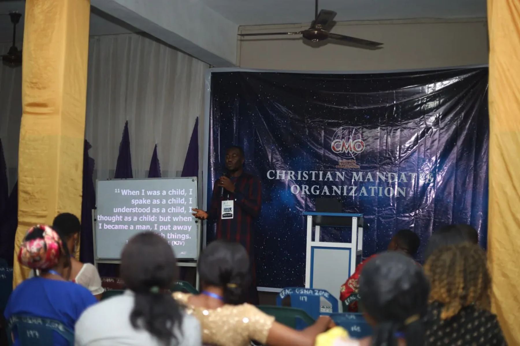 A speaker teaching beside a screen of scripture, in front of the CMO banner