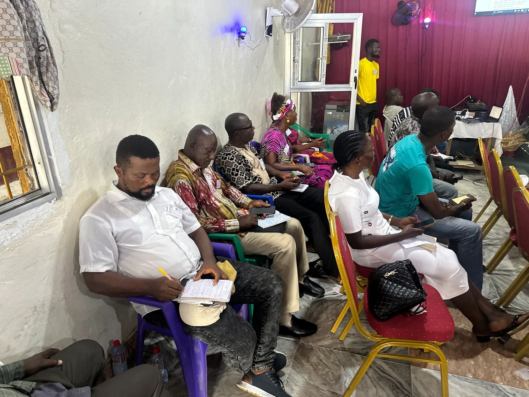 Adults seated along the side of the hall, taking notes