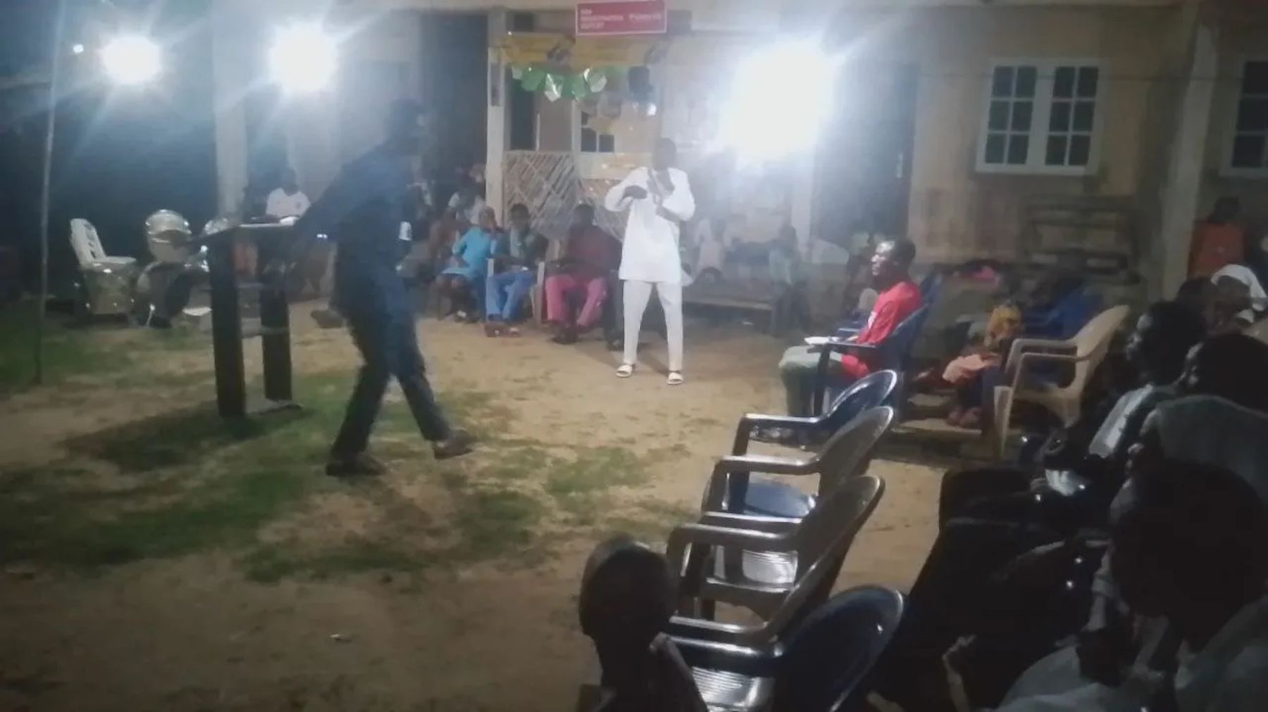 A preacher speaking in an open courtyard at night, with people seated around