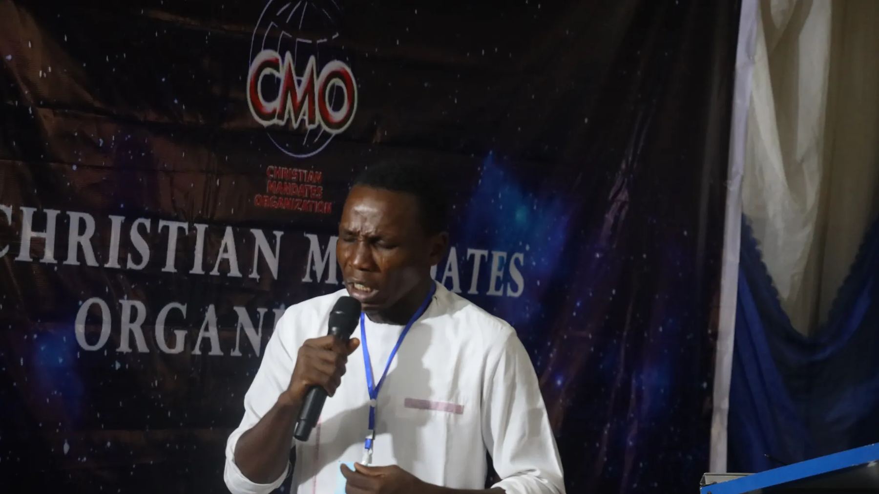 A speaker with a microphone ministering in front of the Christian Mandates Organization banner