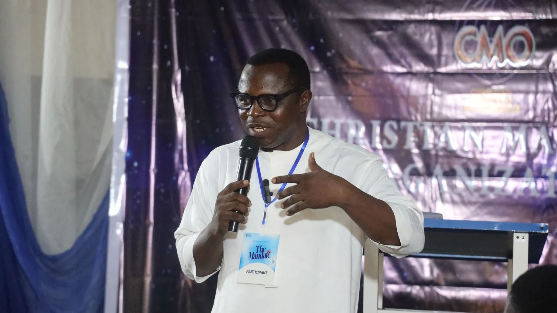 A speaker in glasses teaching with a microphone in front of the CMO banner