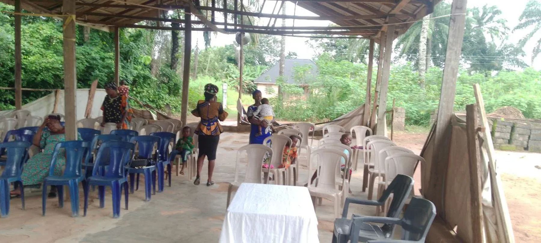 The open-sided church shelter among palm trees, with worshippers gathering on rows of plastic chairs
