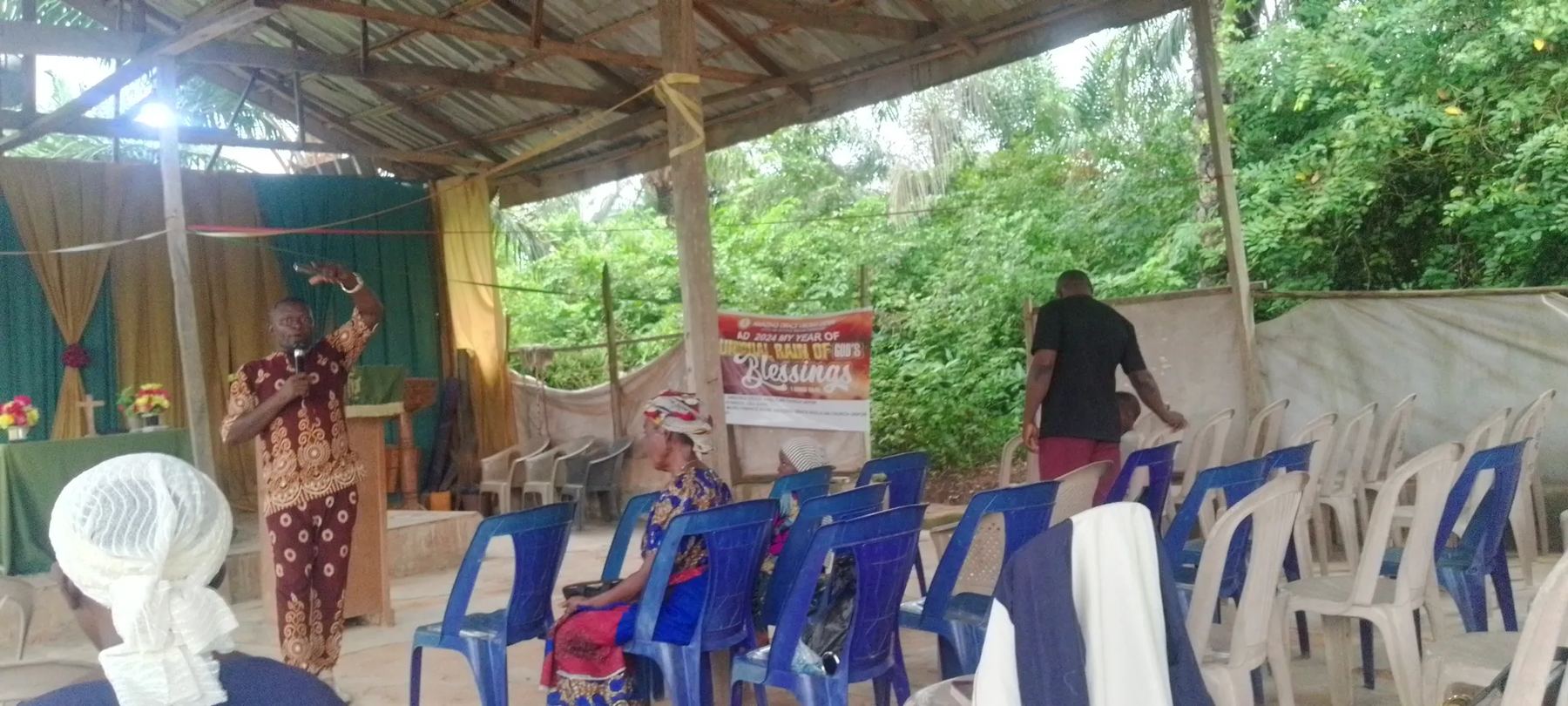 A minister preaching with a raised hand under the open-sided church shelter, with worshippers on blue and white plastic chairs