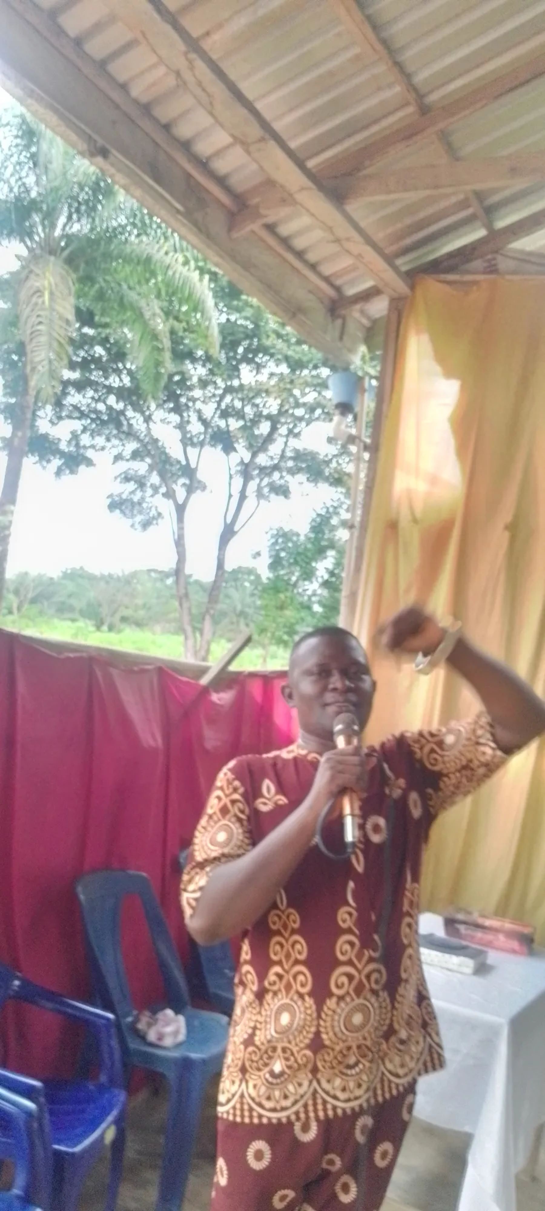 A minister in a patterned outfit preaching with a microphone, one hand raised