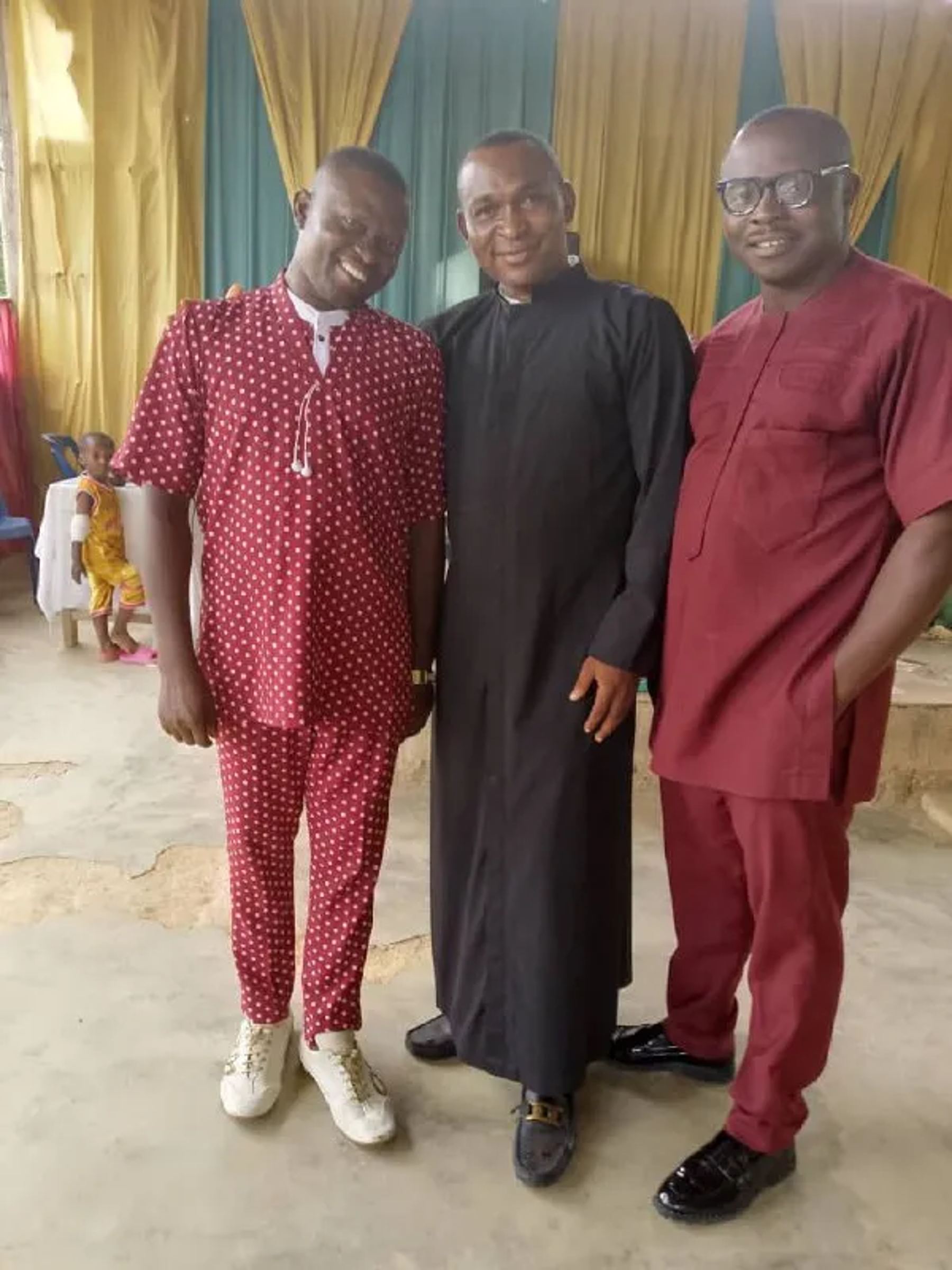 Three ministers, one in a black cassock, standing together in front of the church curtains